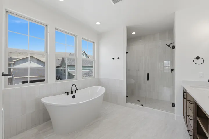 Primary suite. Freestanding soaking tub with a matte black faucet, glass-enclosed shower, dual vanity with wood-finish cabinetry, and large windows