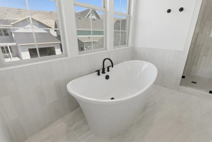 Primary suite. Freestanding soaking tub with matte black fixtures, set against light tile wainscoting and light flooring