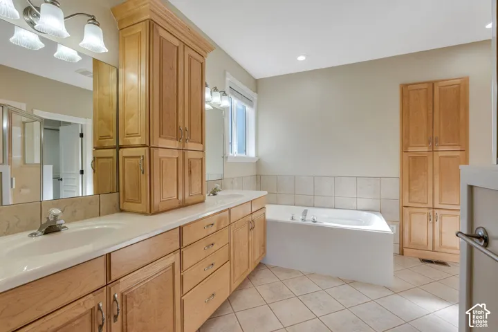 Double vanity with integrated sinks, light wood-finish cabinetry, built-in soaking tub, and tiled flooring