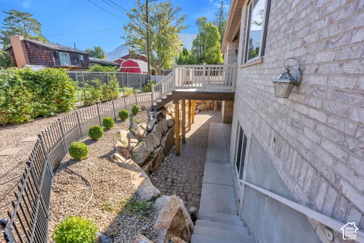 Elevated wood-finish deck with railings, light-colored brick exterior, paver patio, stone retaining wall, and wrought iron fence
