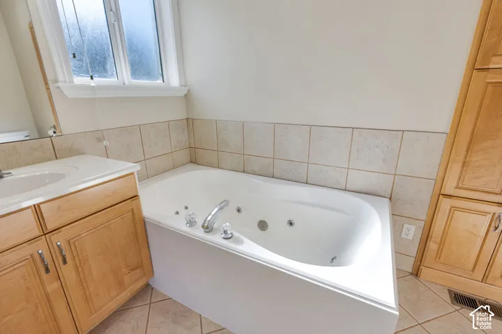 Corner soaking tub with jets, light wood-finish vanity with an integrated sink, tiled flooring, and light-toned wall tiling