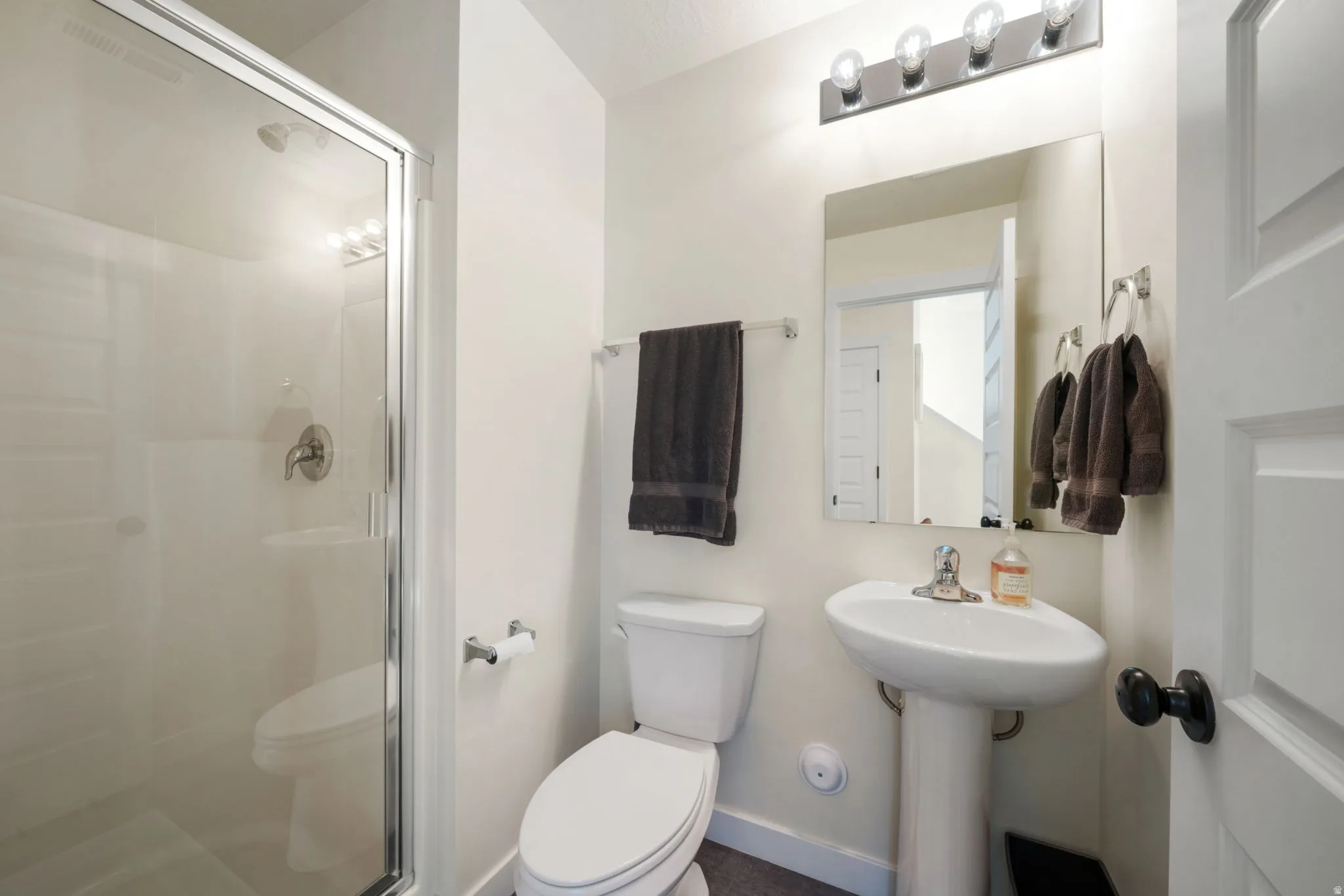 Pedestal sink, wall-mounted mirror, four-bulb vanity light, and a toilet with a shower enclosure