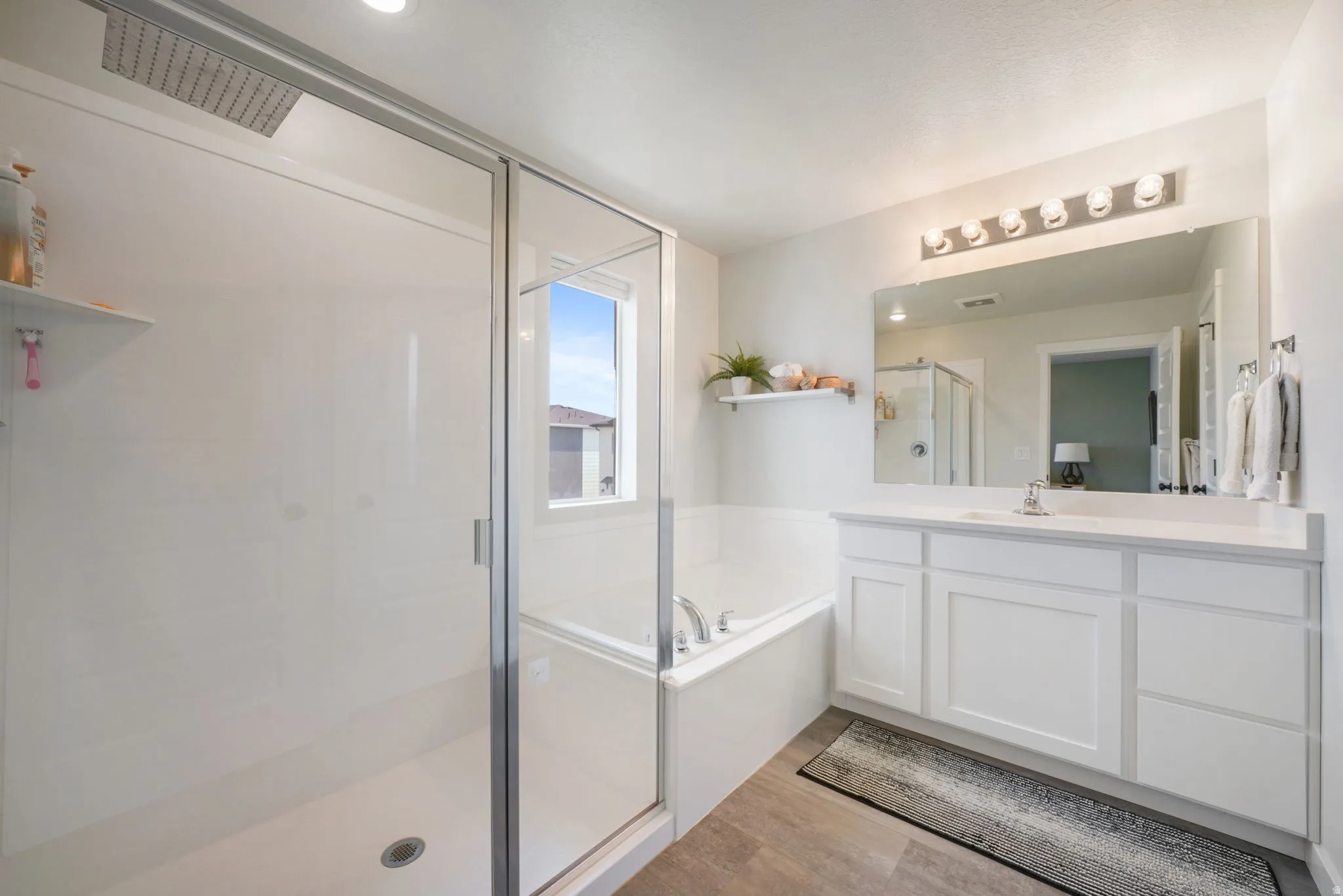 Glass-enclosed shower with a rainfall showerhead, separate soaking tub, double vanity with white cabinetry, and a large mirror with overhead lighting