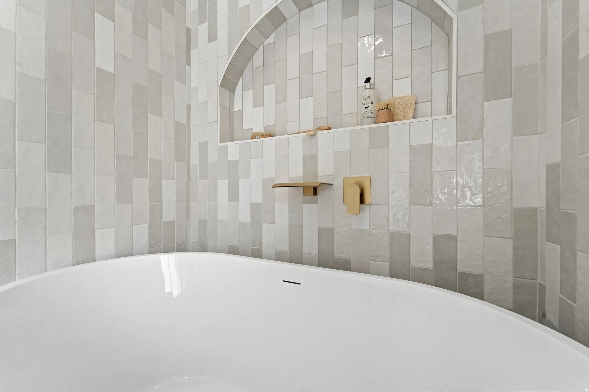 Freestanding soaking tub with gold-tone fixtures, custom tiled niche, and vertical tile surround