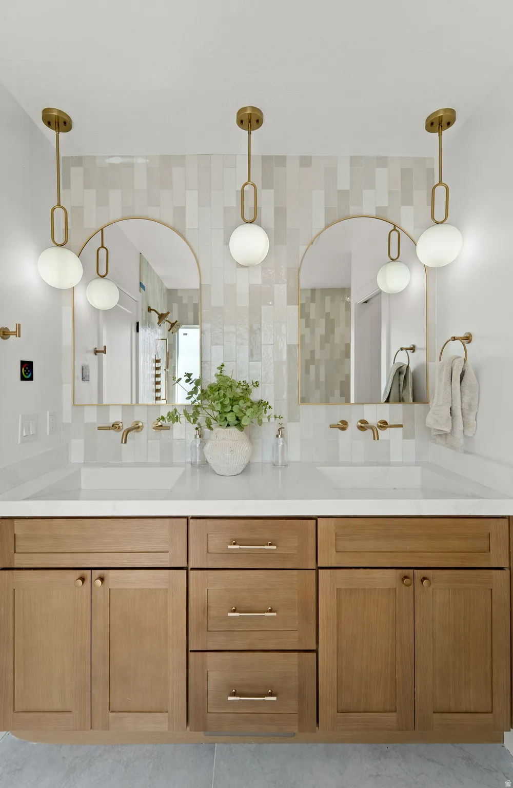 Dual arched mirrors, gold-tone hardware, wood-finish cabinetry, light-toned countertop, and a tiled backsplash