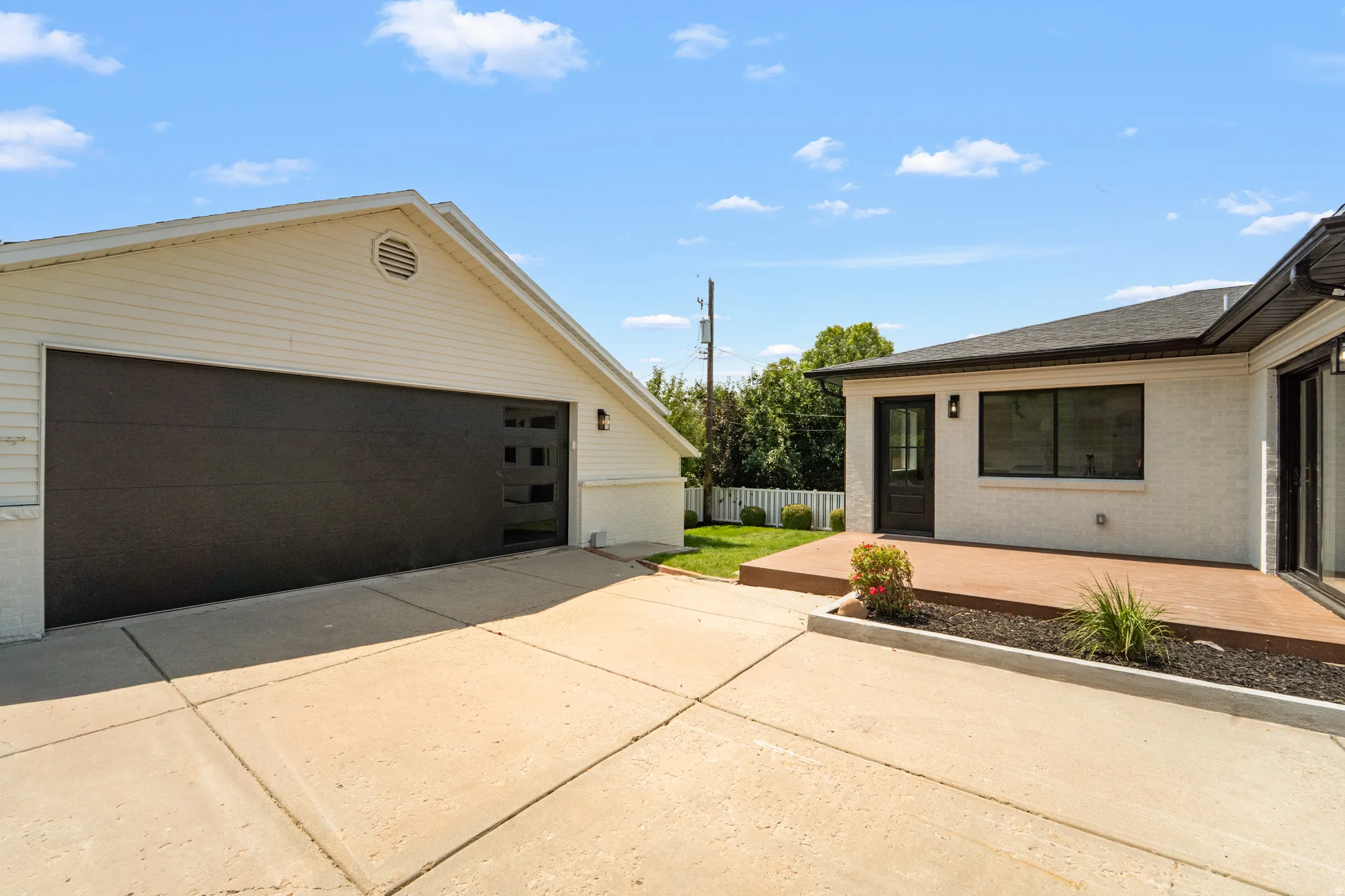 Spacious concrete driveway, modern dark-toned garage door with window inserts, white siding, exterior lighting fixtures, and a wood-finish patio