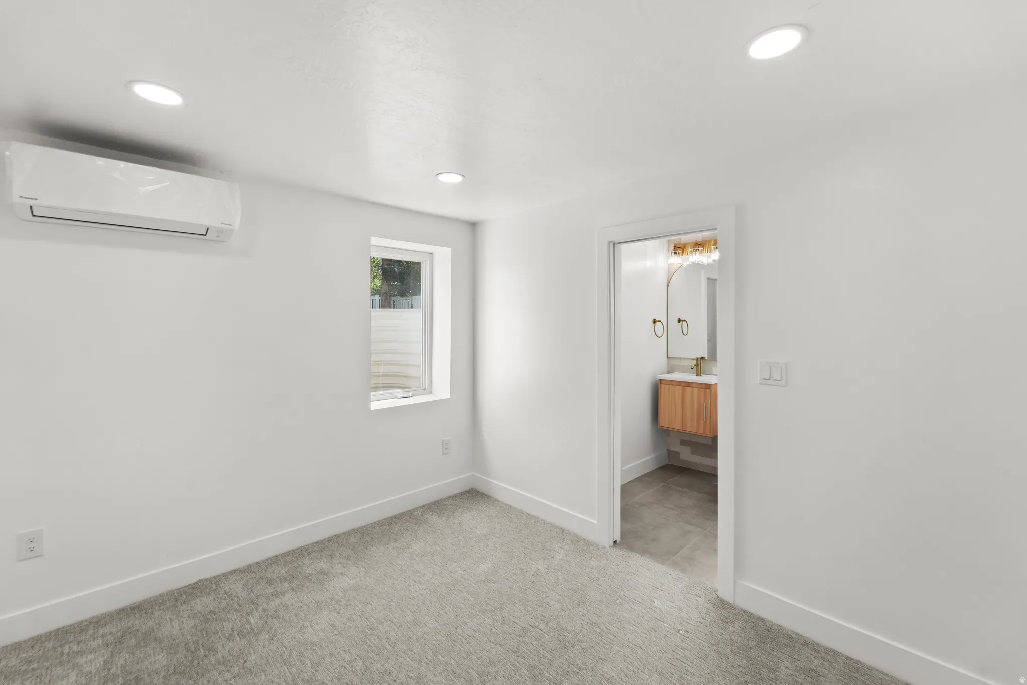 Recessed ceiling lighting, wall-mounted air conditioning unit, gray carpet flooring, and a window with natural light