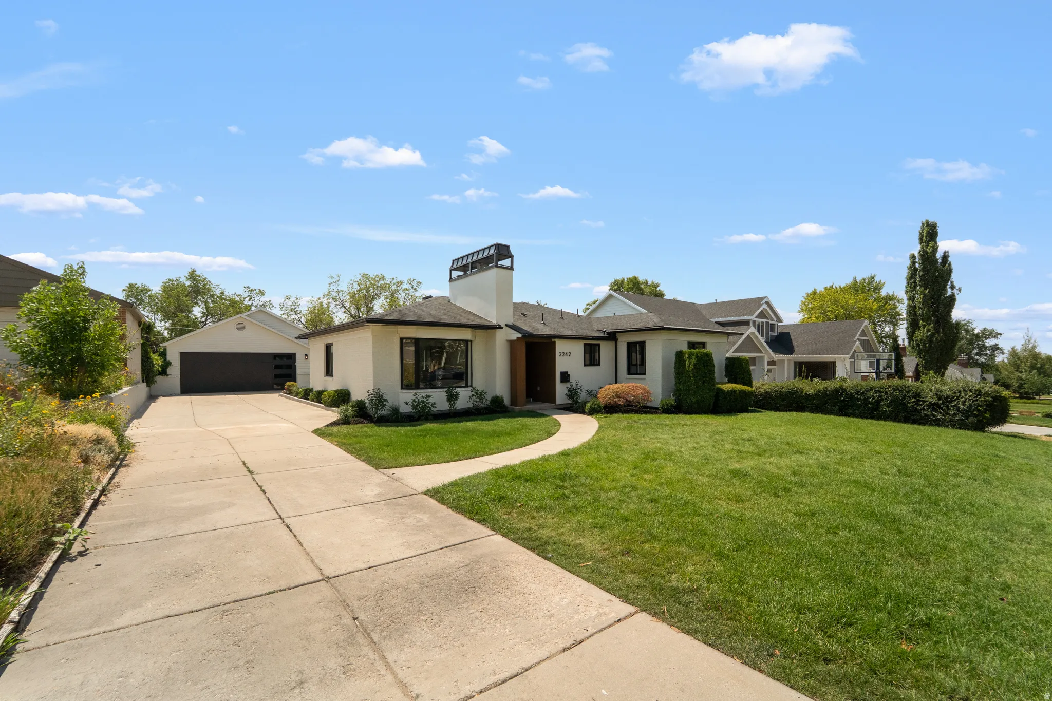 White stucco exterior with dark trim windows, an attached two-car garage, an elongated concrete driveway, a curved pathway, and a front lawn