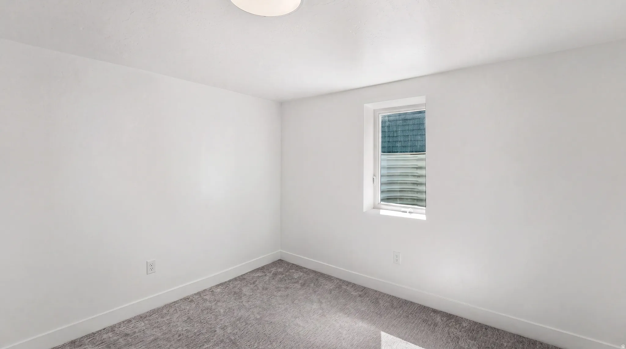 White painted walls with a single window, overhead flush-mount lighting, and gray carpet flooring