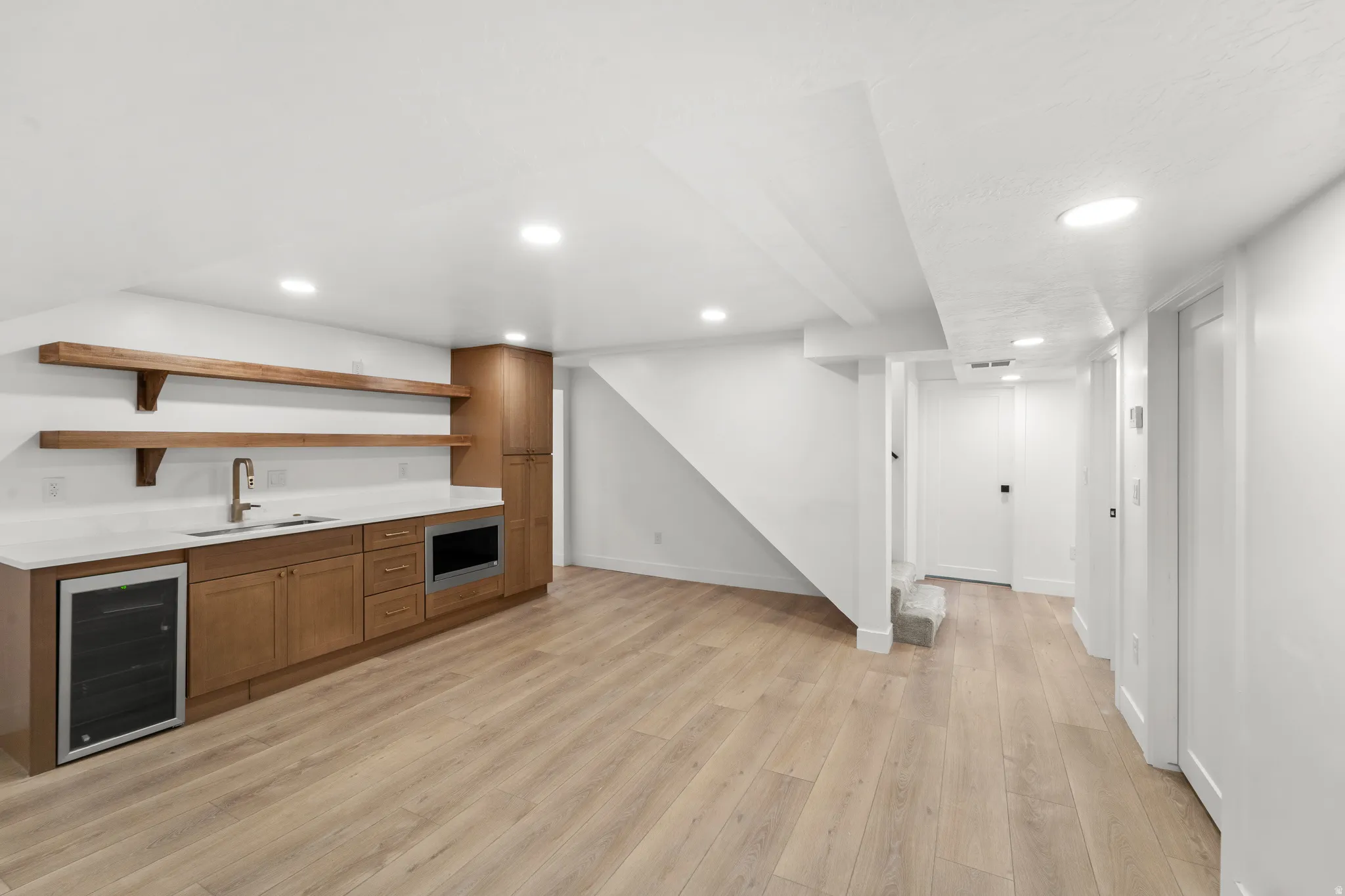 Modern basement entertaining space featuring custom wood cabinetry with integrated appliances, floating wood shelving, wood-finish flooring throughout, recessed ceiling lighting, and open layout with staircase access