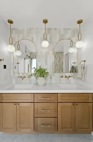 Dual arched mirrors, gold-tone hardware, wood-finish cabinetry, light-toned countertop, and a tiled backsplash