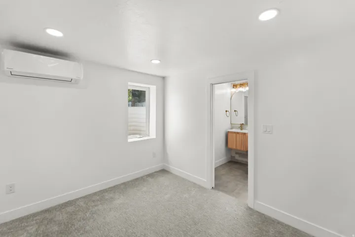 Recessed ceiling lighting, wall-mounted air conditioning unit, gray carpet flooring, and a window with natural light