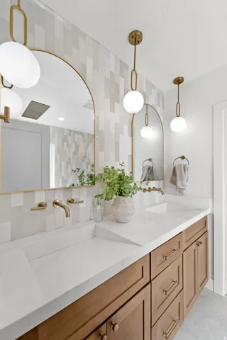Dual arched mirrors, white countertop with integrated sinks, light wood-finish cabinetry, gold-tone hardware, and patterned wall tiling