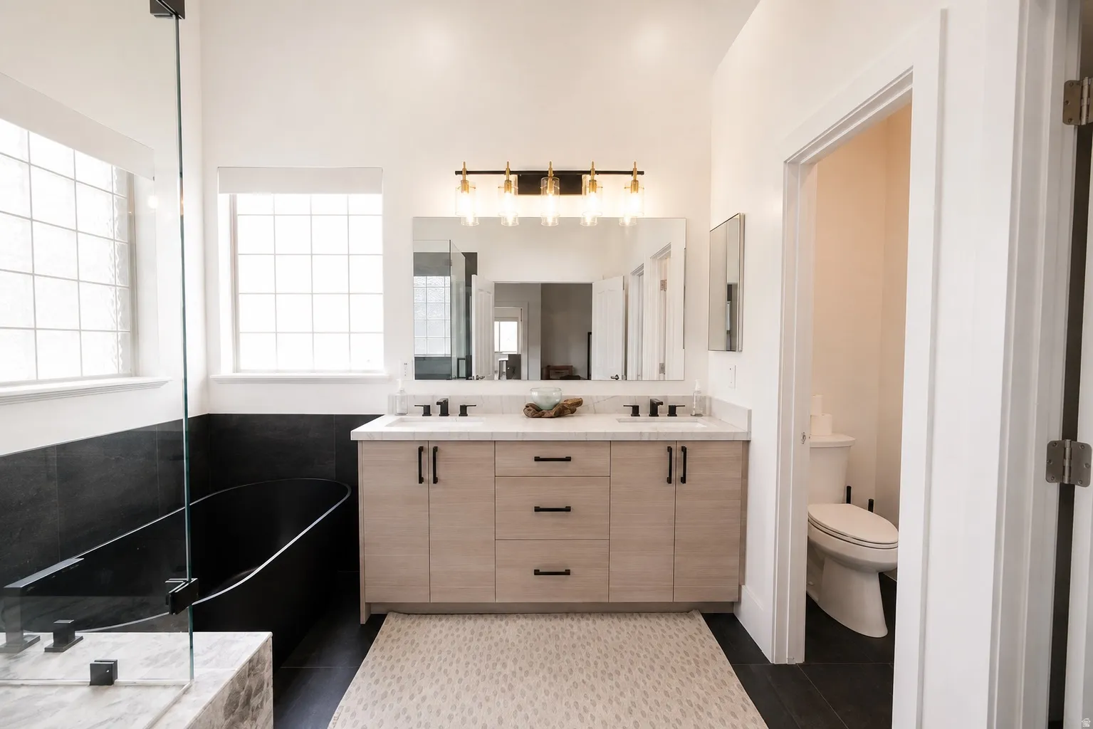 Dual-sink vanity with light-toned wood-finish cabinetry, black hardware, and a light-toned countertop