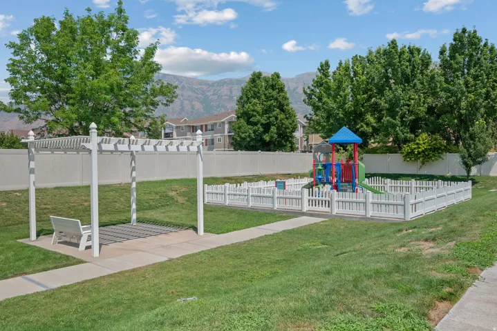 Community green space featuring a pergola with a bench, a paved walkway, a fenced playground, and mature trees