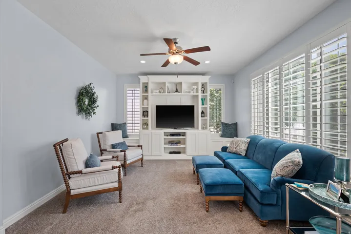 Built-in cabinetry with integrated media center, wood-finish ceiling fan, recessed lighting, expansive windows with plantation shutters, and light-toned carpeting