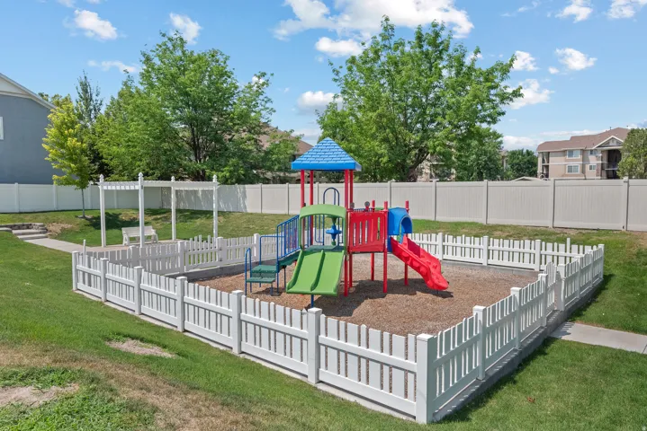 Fenced playground area featuring a colorful play structure with slides, surrounded by a white picket fence and a larger privacy fence