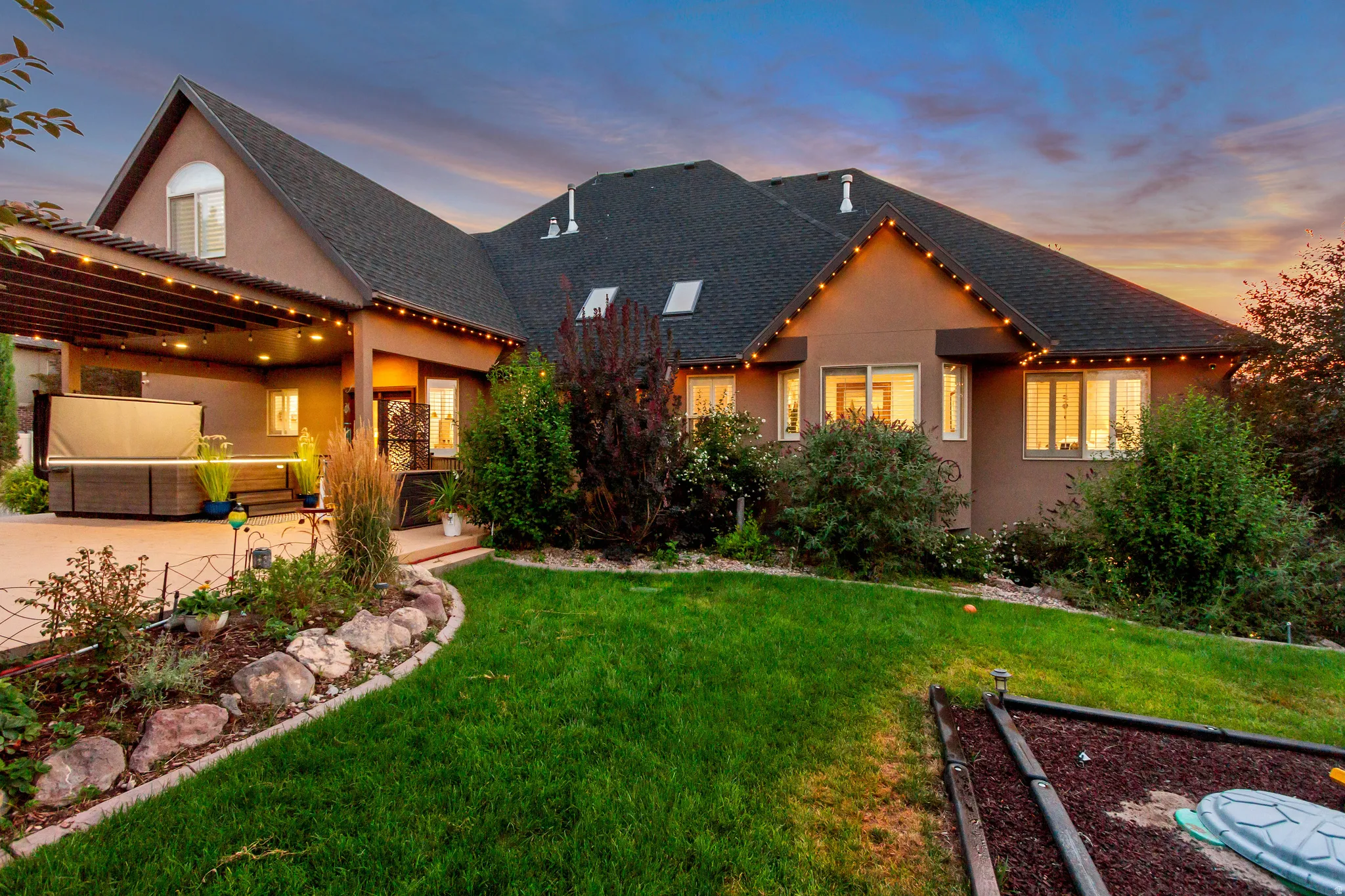 Expansive stucco exterior with multiple windows, a covered patio, and a landscaped yard featuring a rock garden border