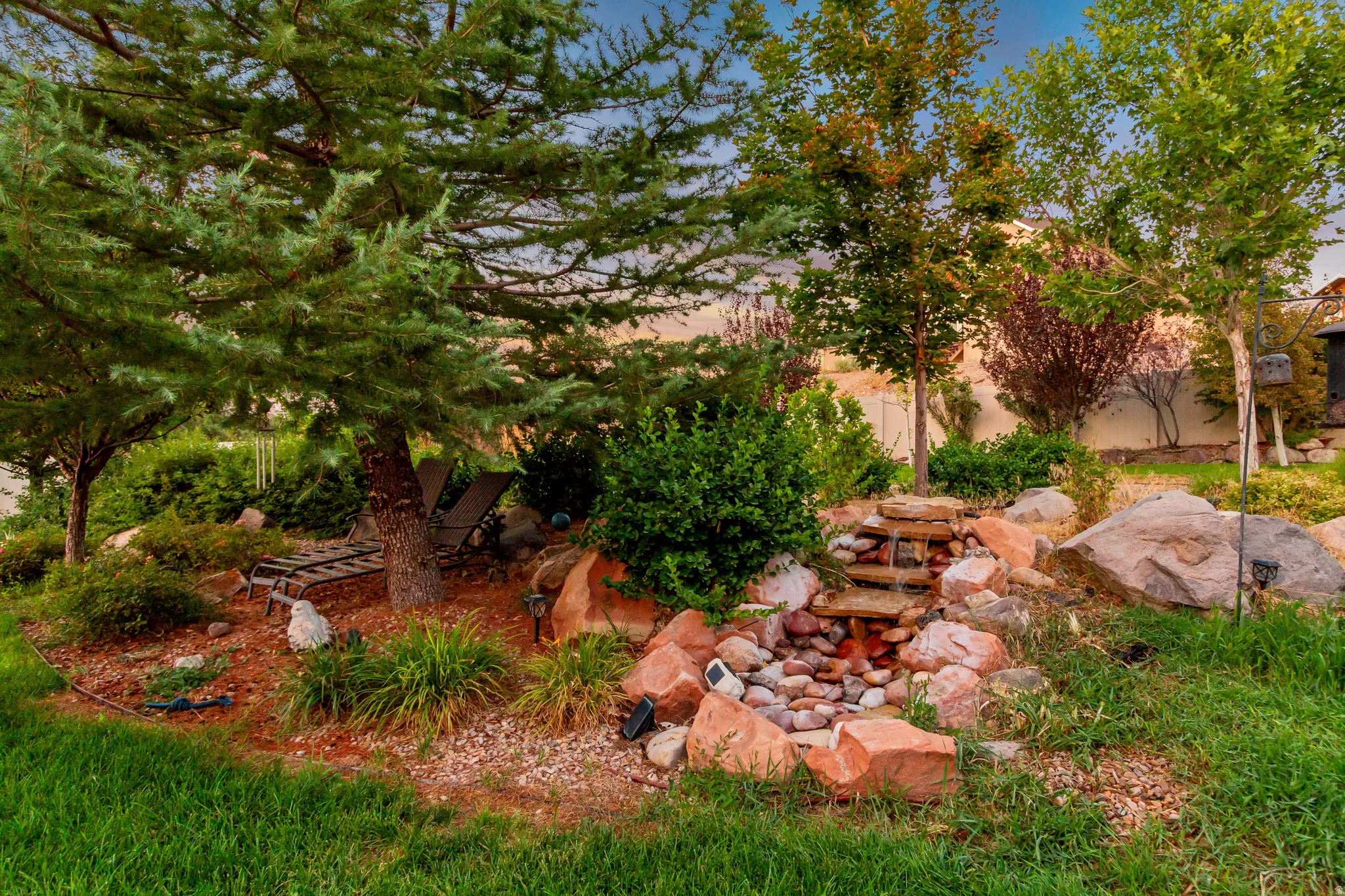 Lush landscaping featuring mature trees, a multi-tiered rock waterfall, decorative boulders, and planted garden beds