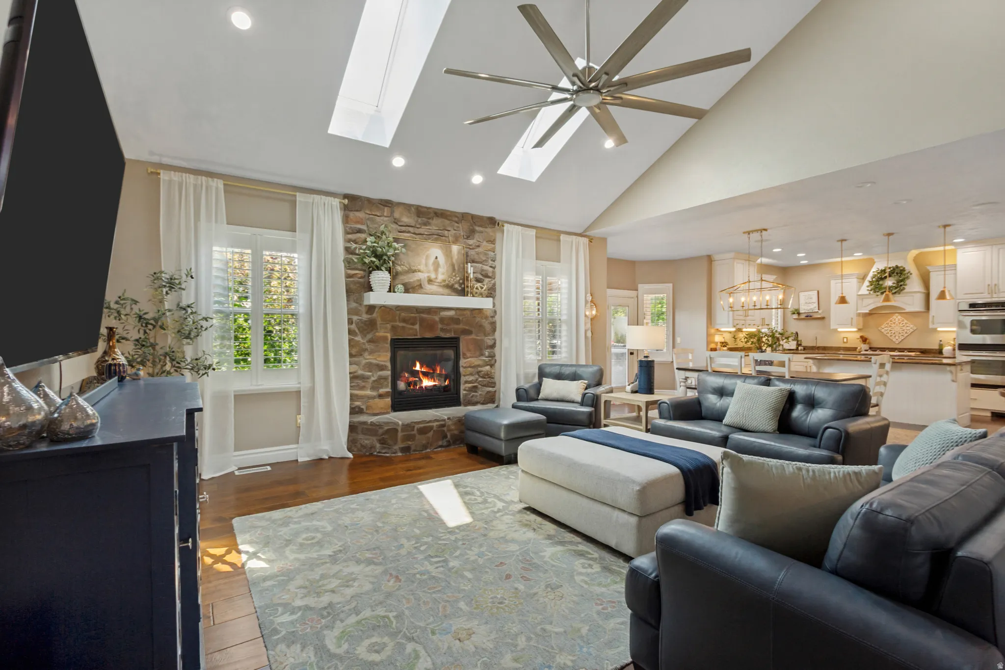 Vaulted ceiling with skylights, wood-finish flooring, stone-faced fireplace with a white mantel, recessed lighting, and a contemporary ceiling fan