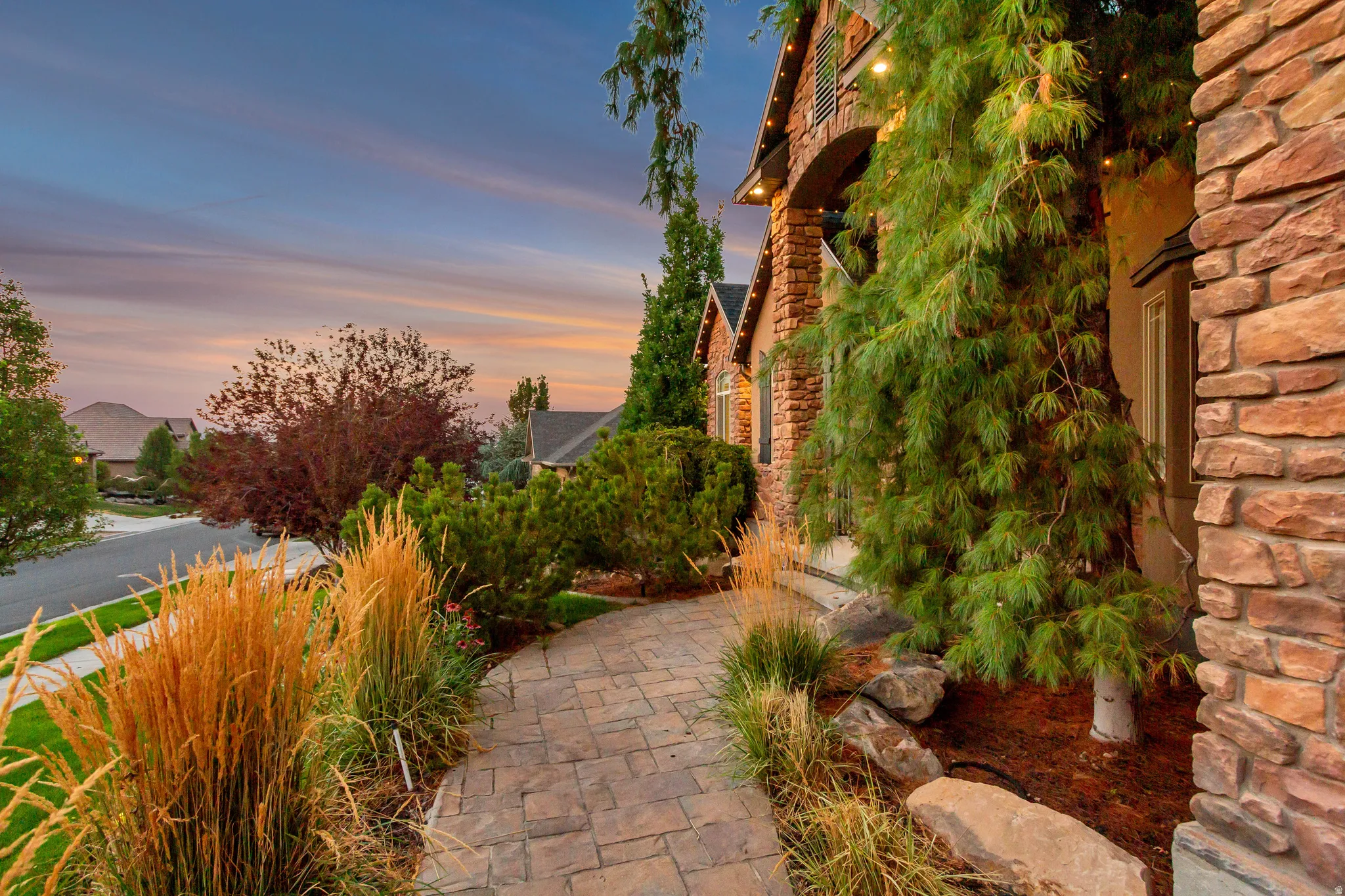 Stone-clad exterior with an arched entryway, complemented by a paver pathway and mature landscaping featuring ornamental grasses and evergreen trees