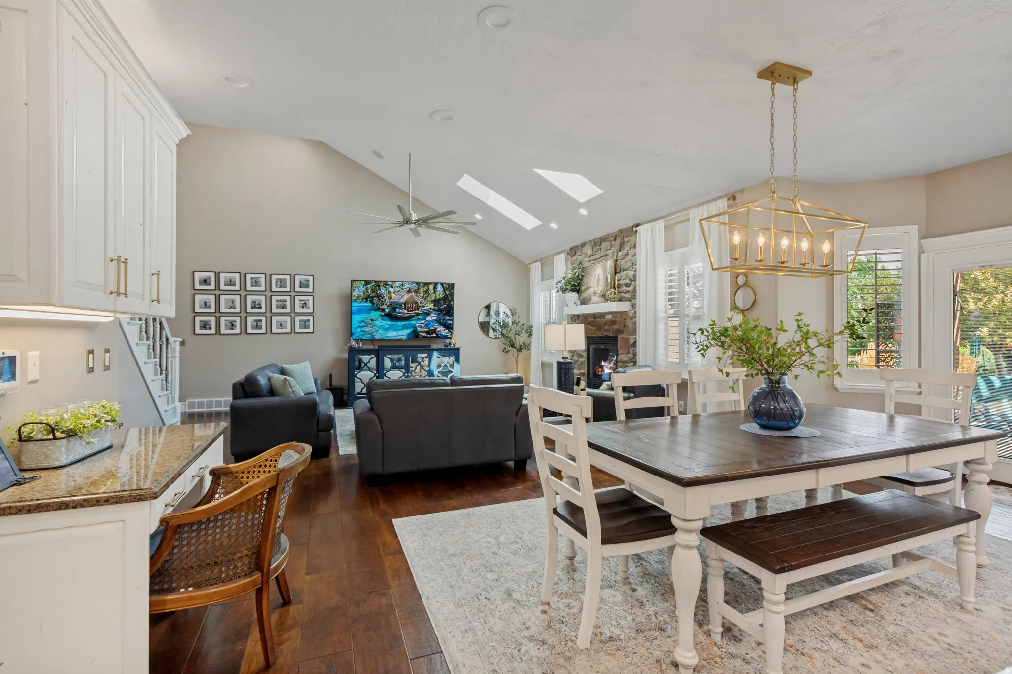 Open-concept living area with vaulted ceilings, skylights, wood-finish flooring, a stone-clad fireplace, and a built-in kitchen desk