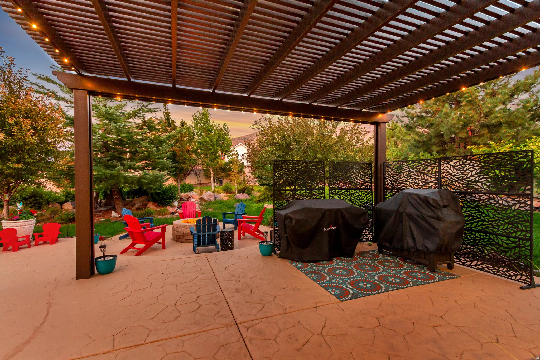 Expansive stamped concrete patio with a louvered pergola, string lighting, decorative privacy screens, and a built-in fire pit