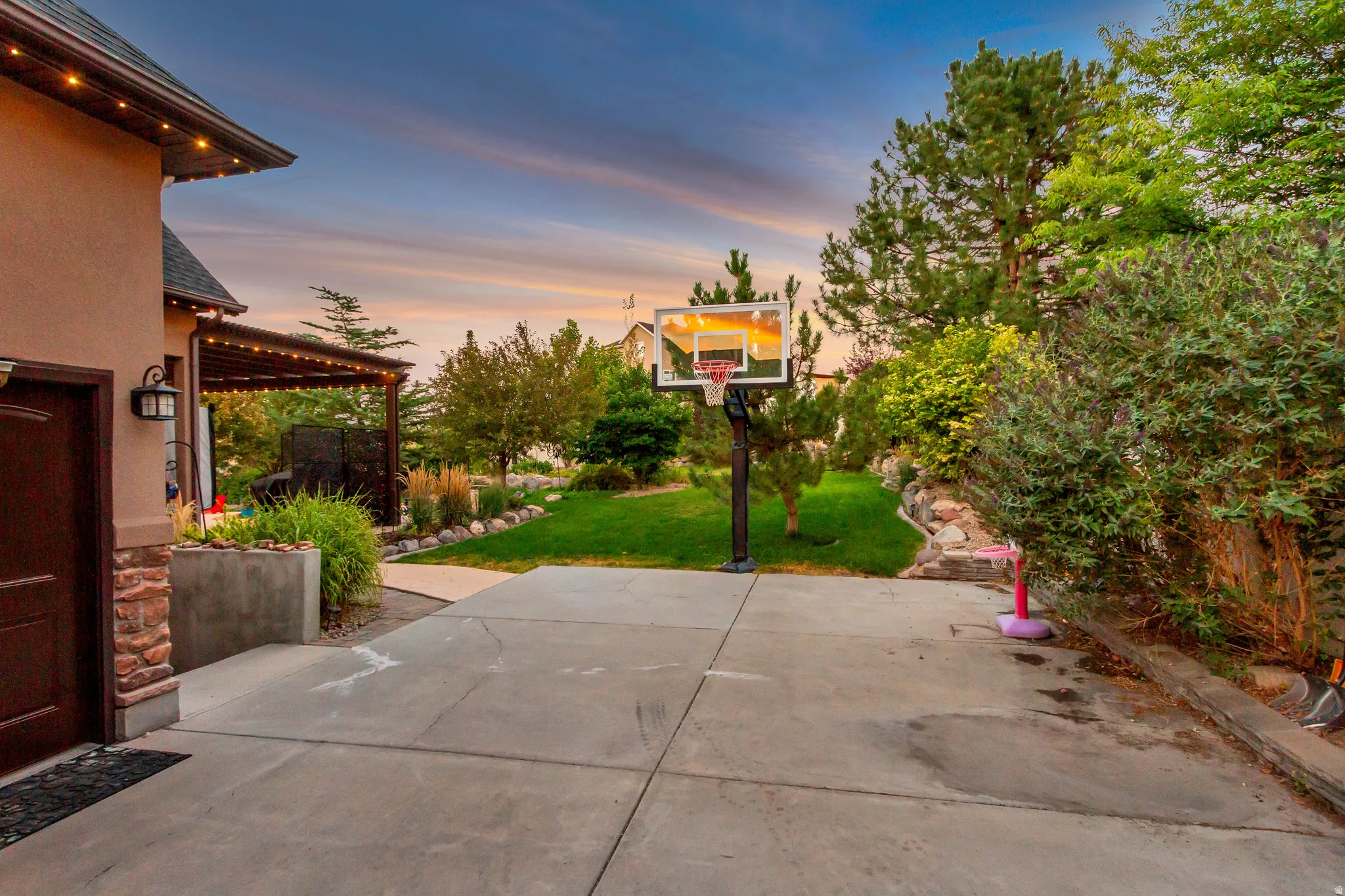 Stucco exterior with stone accents, covered patio with string lights, concrete patio surface, established landscaping, and mature trees