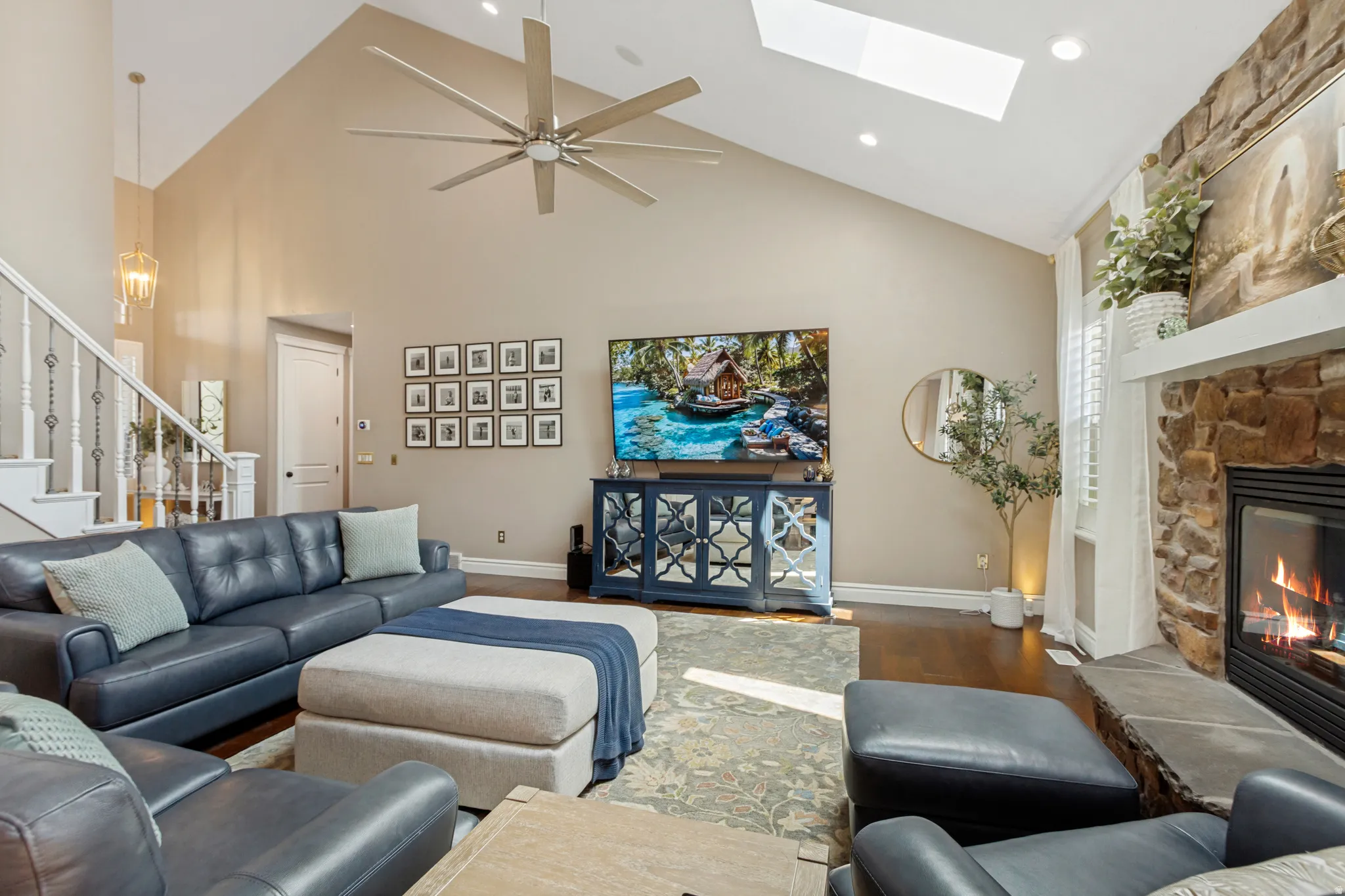 Vaulted ceiling with skylight and ceiling fan, stone fireplace with a white mantel, hardwood flooring, and a white staircase with decorative balusters