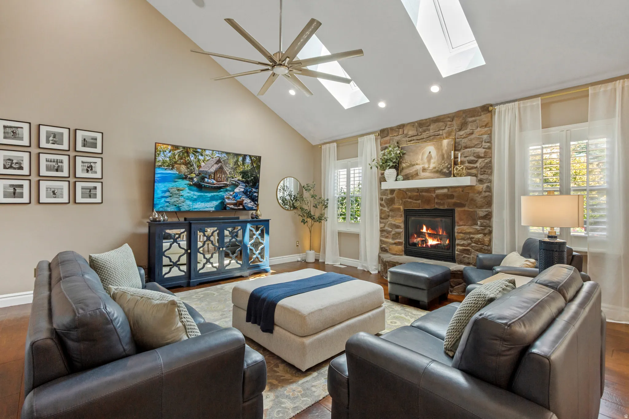 Vaulted ceiling with skylights and an overhead fan, stone-faced fireplace with a white mantel, and wood-finish flooring
