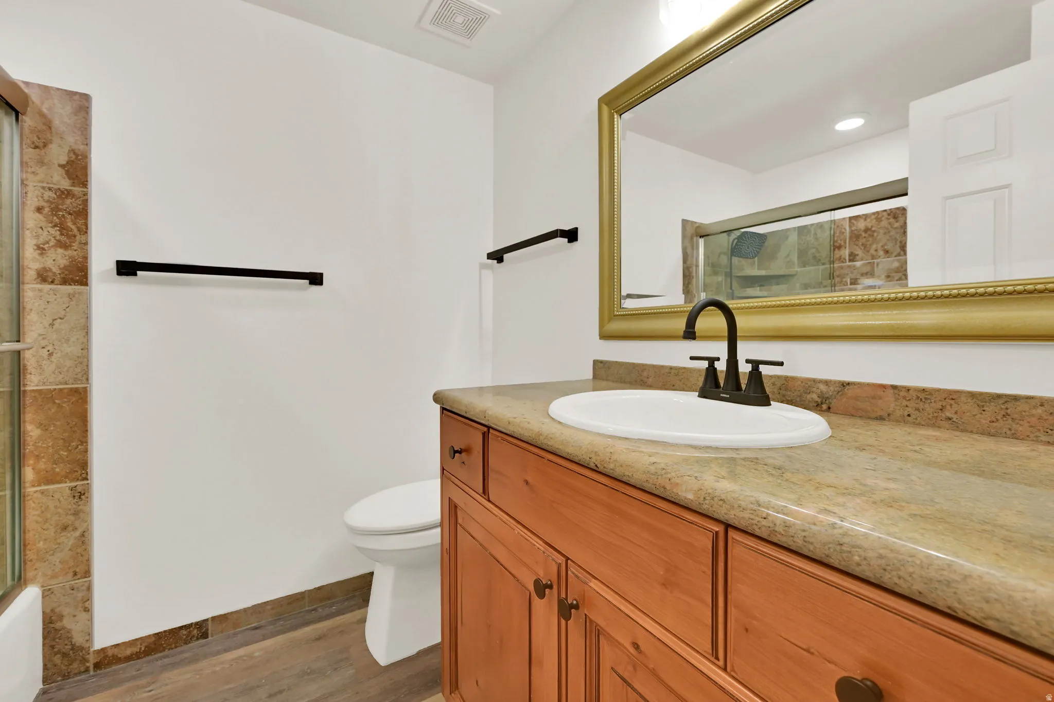 Wood-finish flooring, wood cabinetry, built-in vanity with integrated sink, gold-framed mirror, and tiled shower enclosure