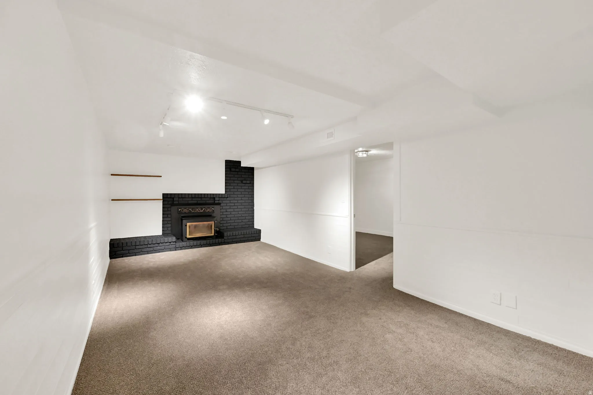 Recessed fireplace with a black brick surround and hearth, track lighting, two built-in floating shelves, and neutral carpet flooring