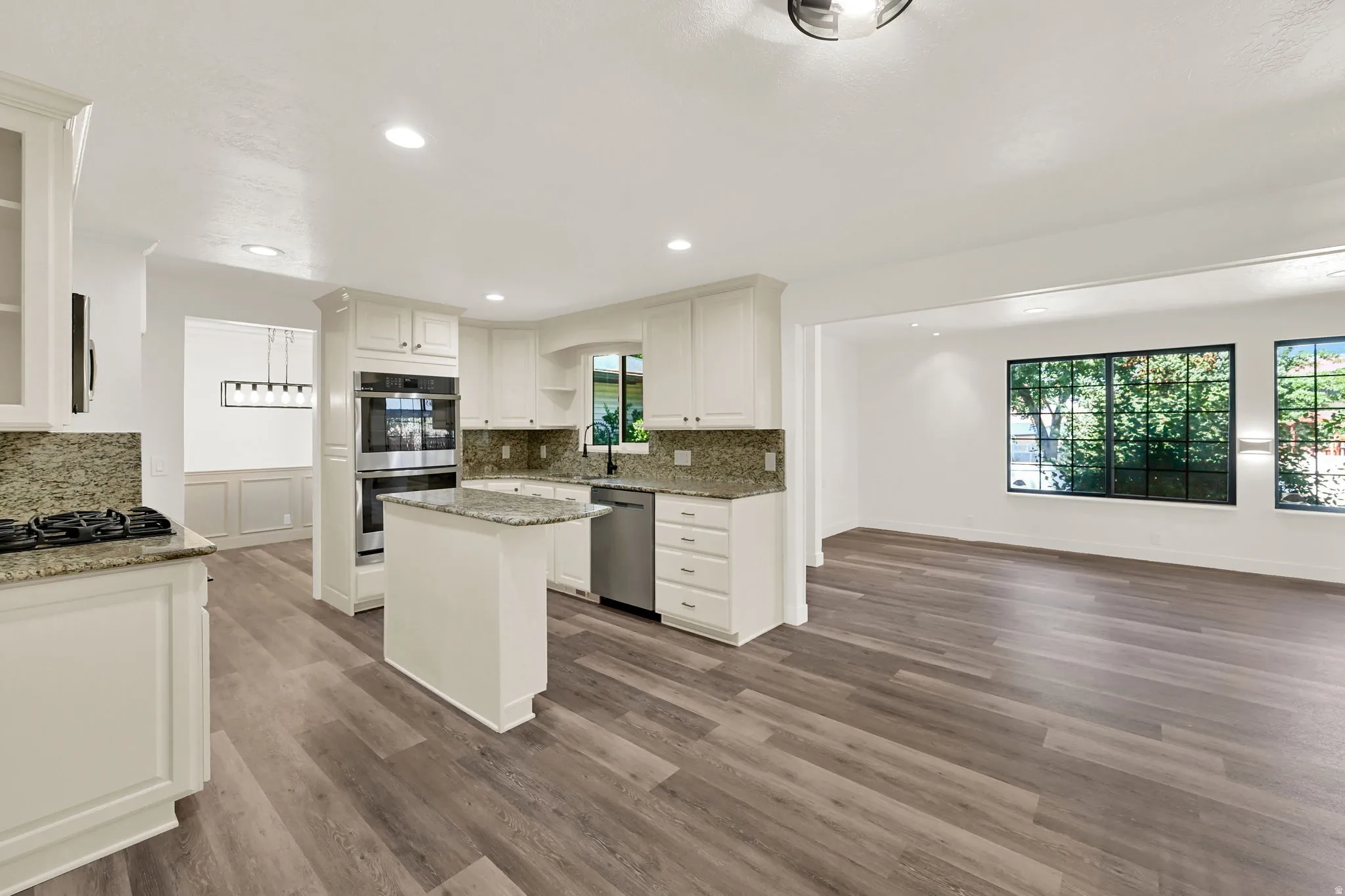 Kitchen featuring white cabinetry, granite countertops, a central island, stainless steel double ovens, and wood-finish flooring extending into an adjacent living area with multiple windows