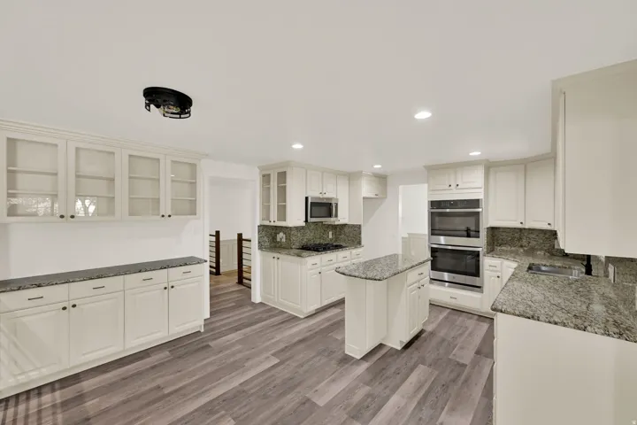 White cabinetry with dark hardware, stone countertops with matching backsplash, stainless steel double ovens, central kitchen island, and wood-finish flooring