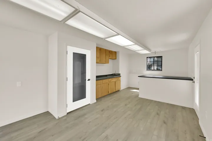 Wood-finish flooring throughout, integrated ceiling light panels, light wood cabinetry, dark countertop, and a glass-paneled door
