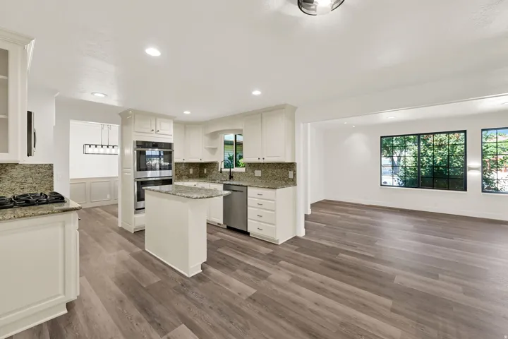 Kitchen featuring white cabinetry, granite countertops, a central island, stainless steel double ovens, and wood-finish flooring extending into an adjacent living area with multiple windows