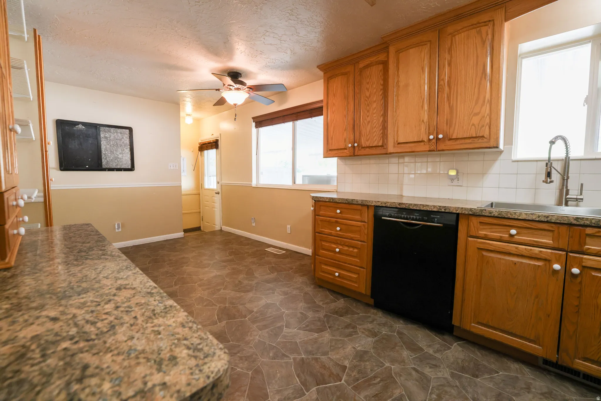 Spacious kitchen featuring wood cabinetry, stainless steel appliances including a black dishwasher, dual sink with chrome faucet, subway tile backsplash, and stone-finish flooring throughout