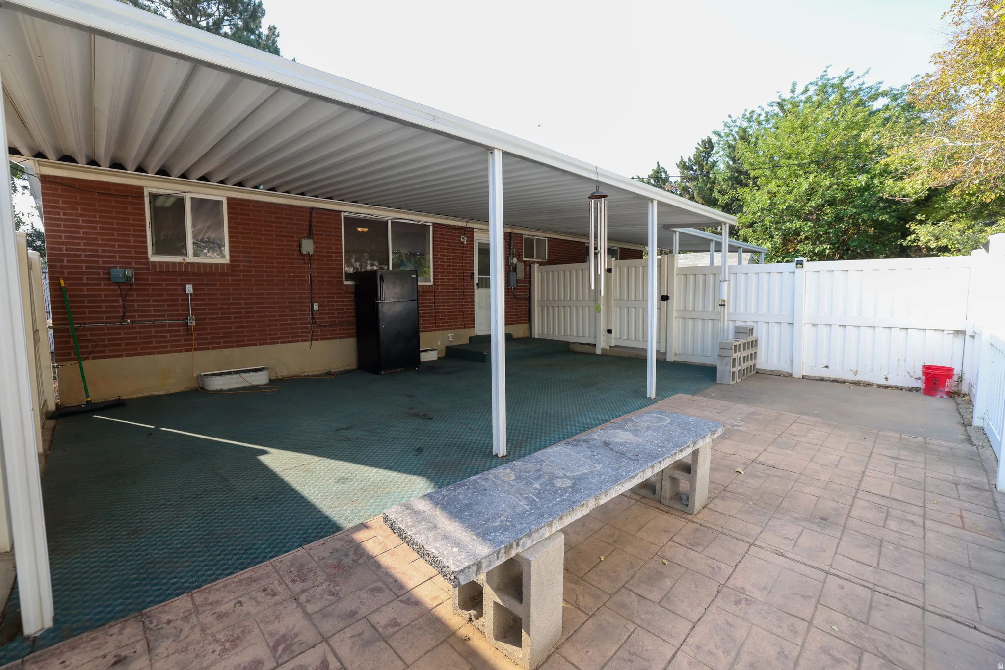 Covered patio area with a brick exterior, concrete pavers, and white privacy fencing