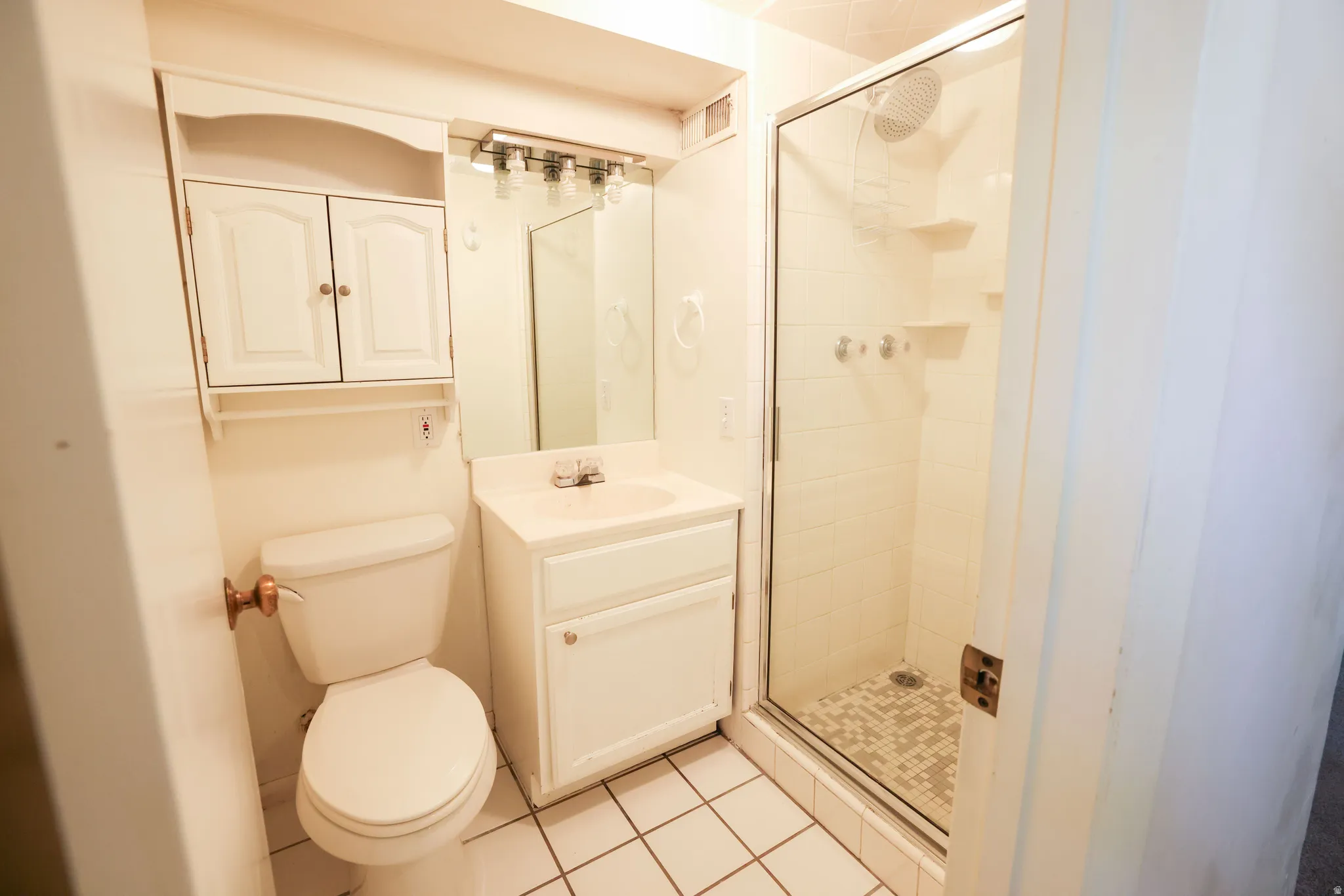White tile flooring with matching wall tile, glass-enclosed shower, white vanity with integrated sink, wall-mounted mirror, and overhead cabinet storage