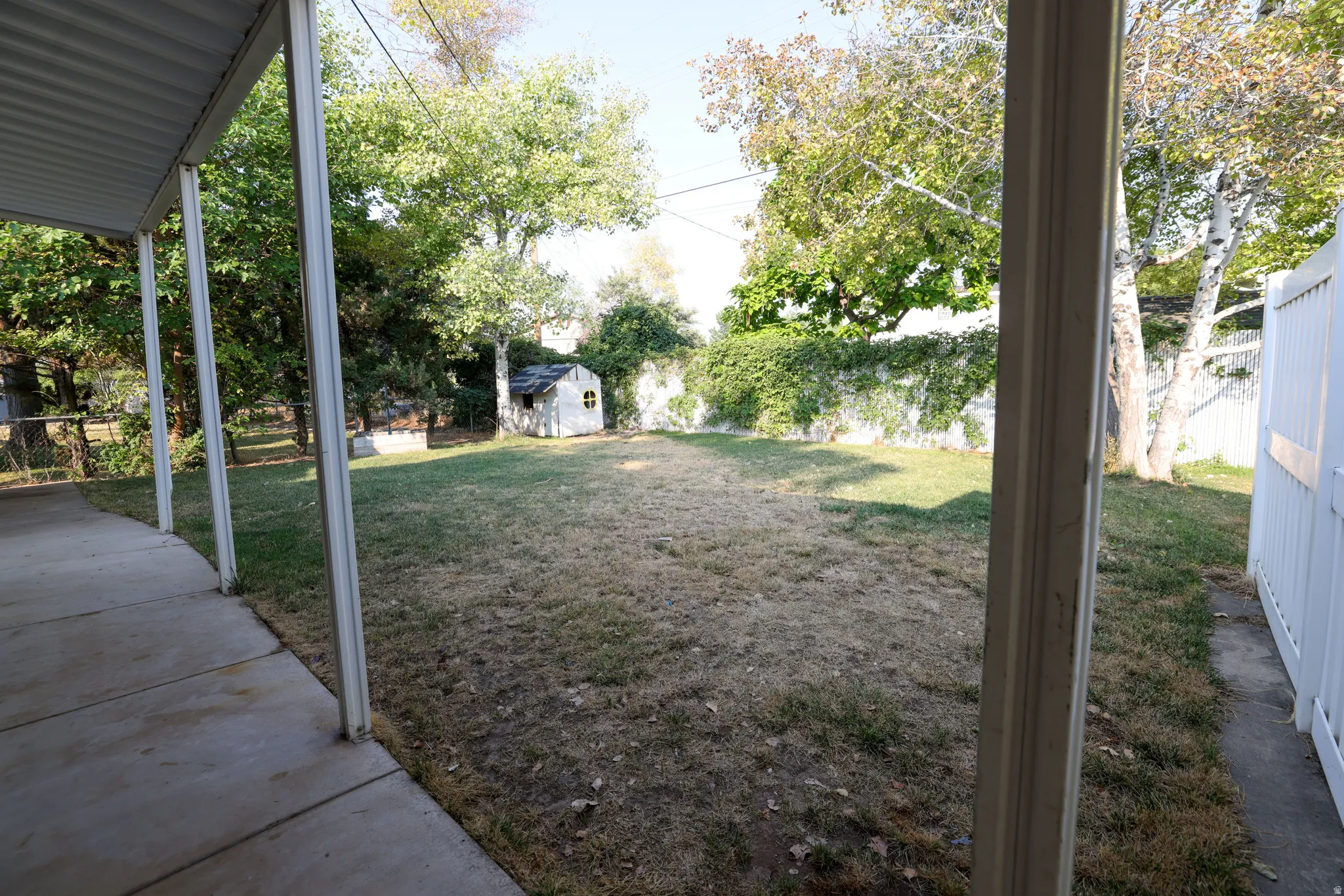 Covered patio with concrete flooring, fenced yard, mature trees, and a storage shed