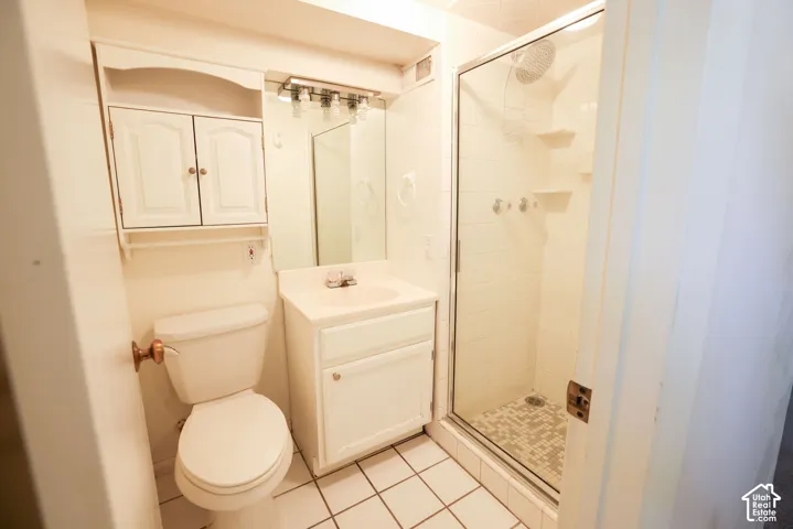 White tile flooring with matching wall tile, glass-enclosed shower, white vanity with integrated sink, wall-mounted mirror, and overhead cabinet storage