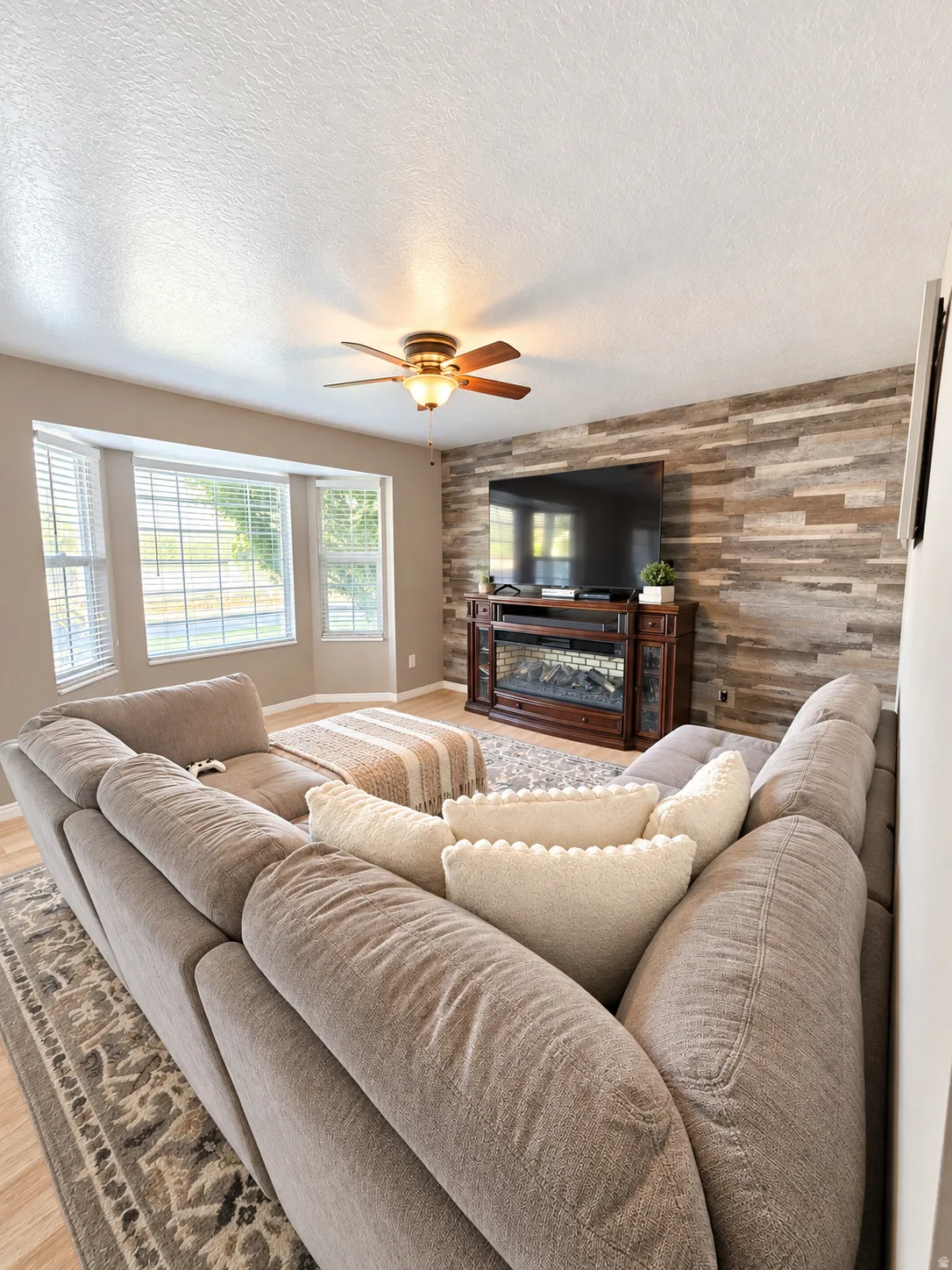 Bay window with blinds, wood-finish flooring, accent wall with horizontal wood paneling, and a ceiling fan with integrated lighting