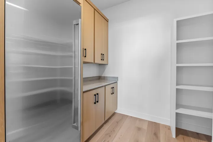 Built-in stainless steel refrigerator, light wood-finish cabinetry with black hardware, light-toned countertop, white built-in shelving, and wood-finish flooring