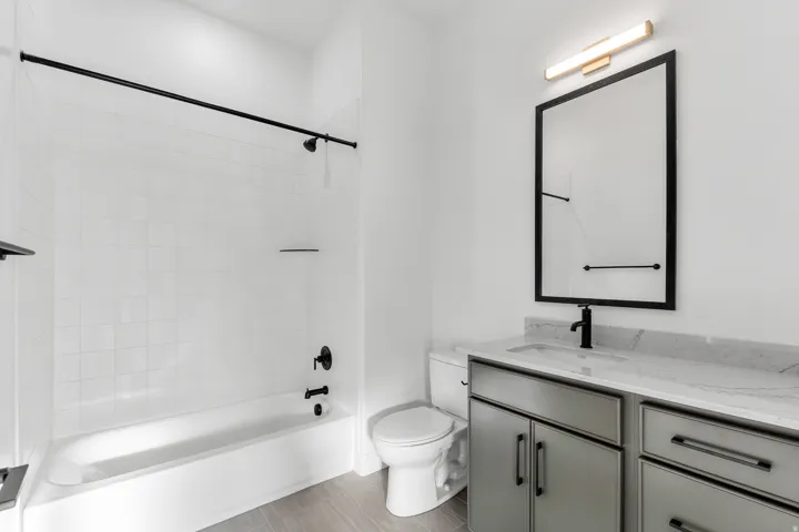 White tiled shower with black matte fixtures, sleek vanity with a light-colored countertop, rectangular undermount sink, and wood-finish flooring
