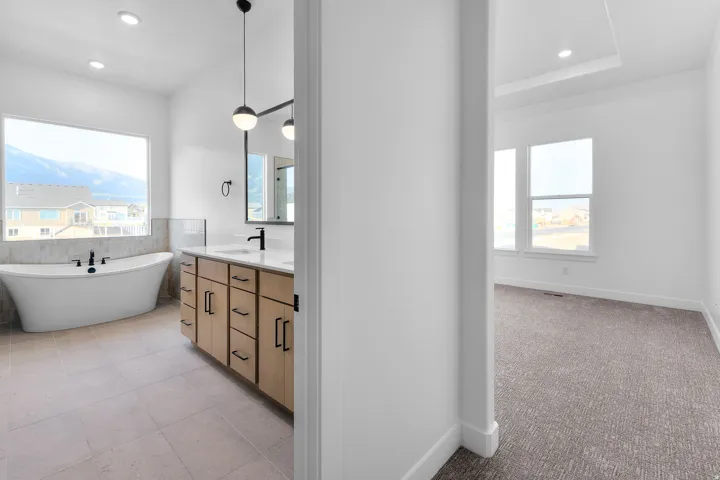 Freestanding soaking tub, double vanity with light wood cabinetry, dual pendant lighting, and large windows