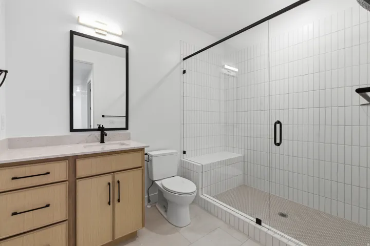 Glass-enclosed shower with white vertical subway tile, built-in shower bench, and dark-framed vanity mirror with integrated lighting