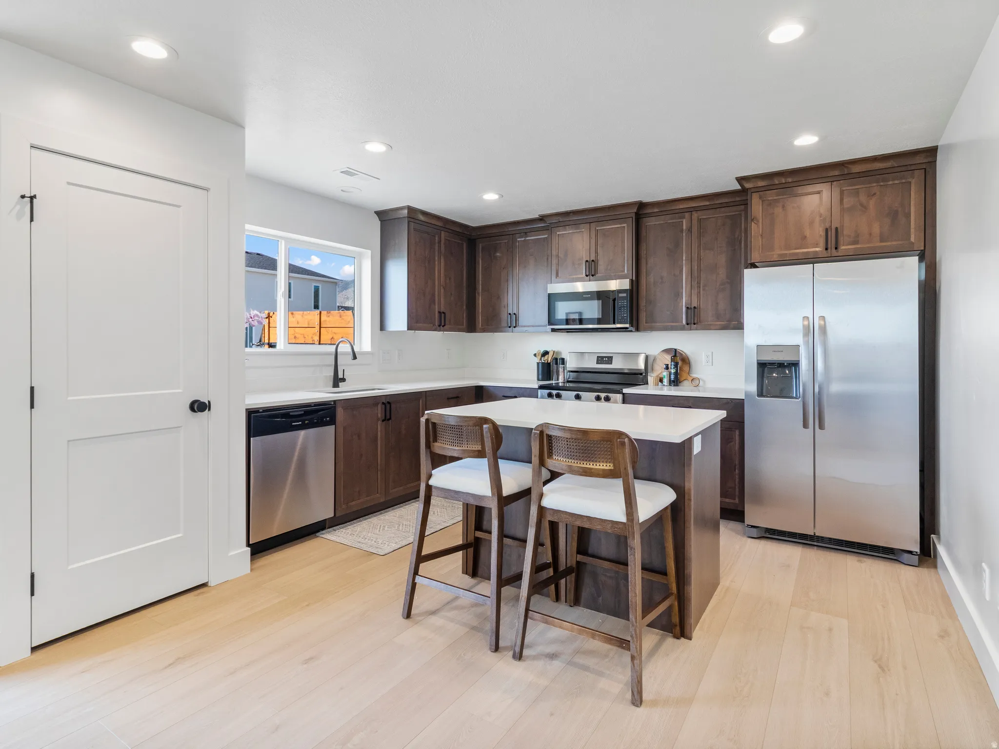 Kitchen featuring wood-finish flooring, recessed lighting, dark wood cabinetry, stainless steel appliances, and a central island with a light-toned countertop