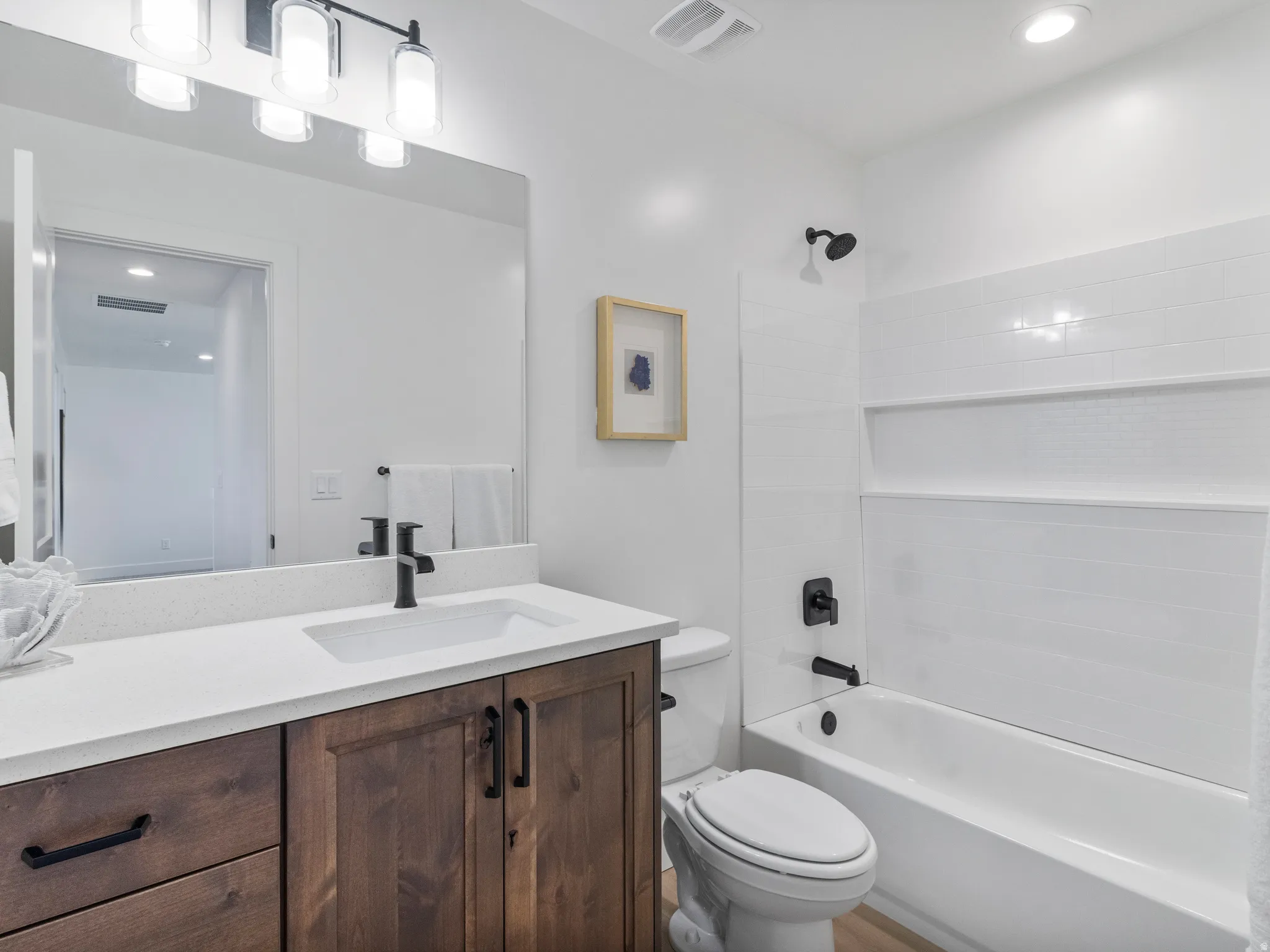 Bathroom featuring a wood-finish vanity with a white countertop, an integrated sink, a rectangular mirror, a four-light fixture, and a tub with subway tile surround and matte black fixtures