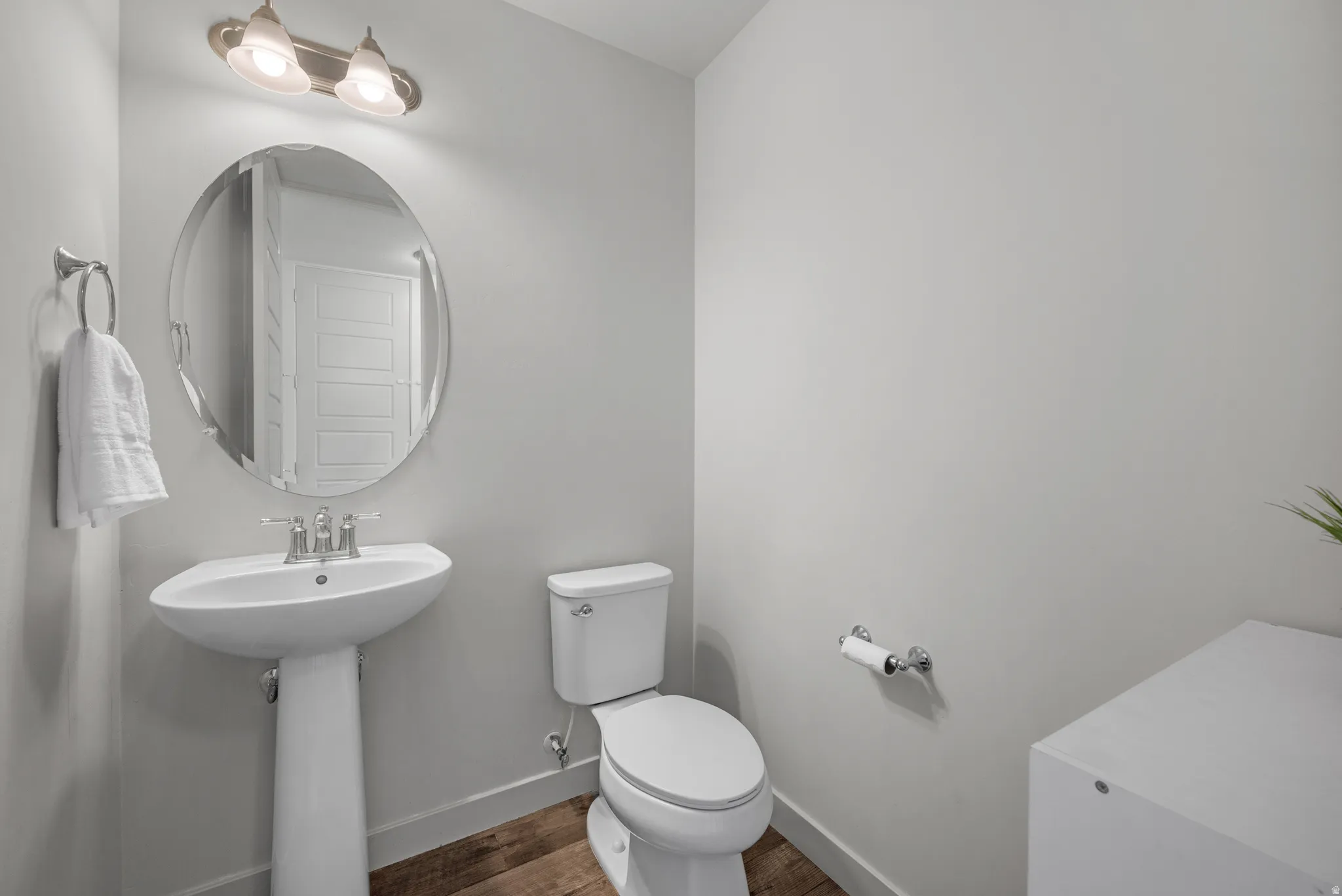 Pedestal sink with chrome faucet, oval mirror, wall-mounted light fixture, white toilet, and wood-finish flooring