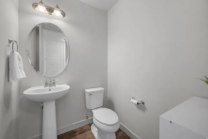 Pedestal sink with chrome faucet, oval mirror, wall-mounted light fixture, white toilet, and wood-finish flooring